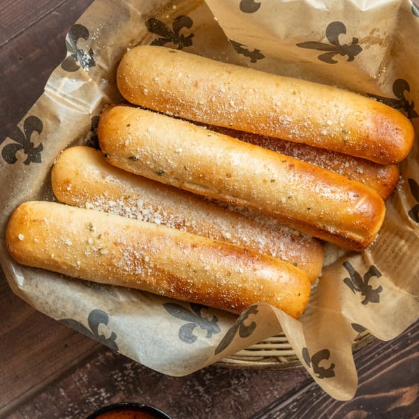 A basket of golden-brown breadsticks, sprinkled with parmesan cheese and herbs, on Alesi Pizza House branded paper.